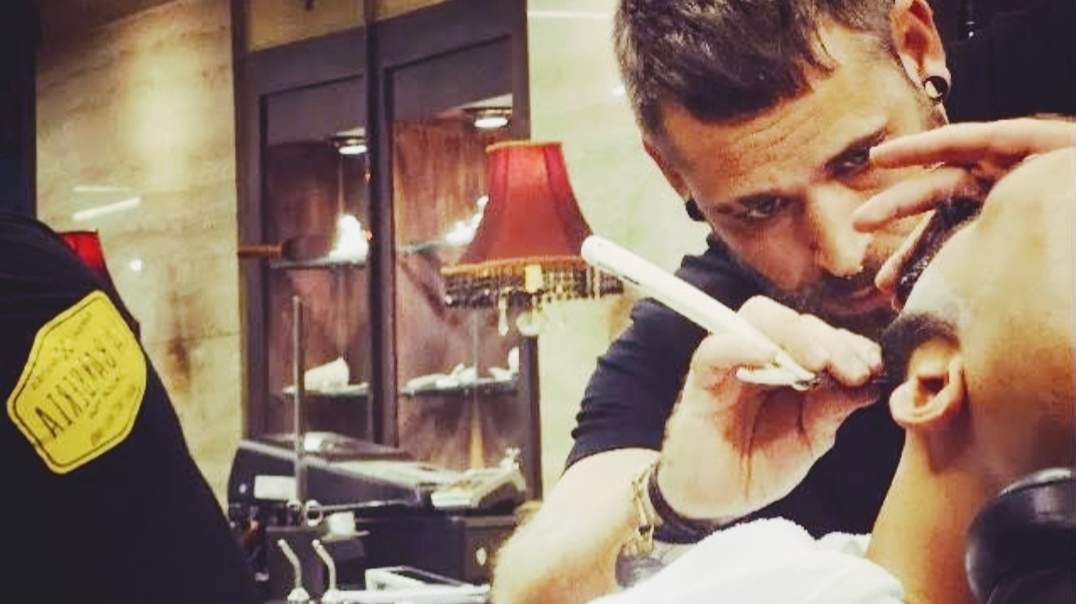 Un barbero perfila con navaja la barba de un cliente, bajo una luz cálida y con una lámpara de flecos roja al fondo.