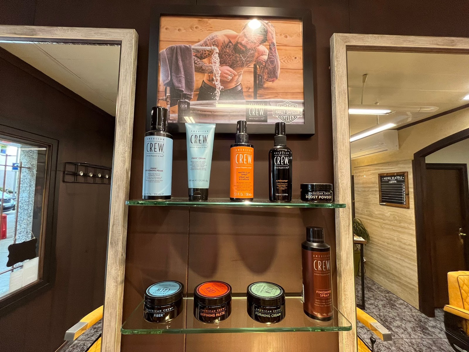 Estantes de cristal con productos de afeitado y peinado bajo un cartel enmarcado, entre dos espejos grandes.