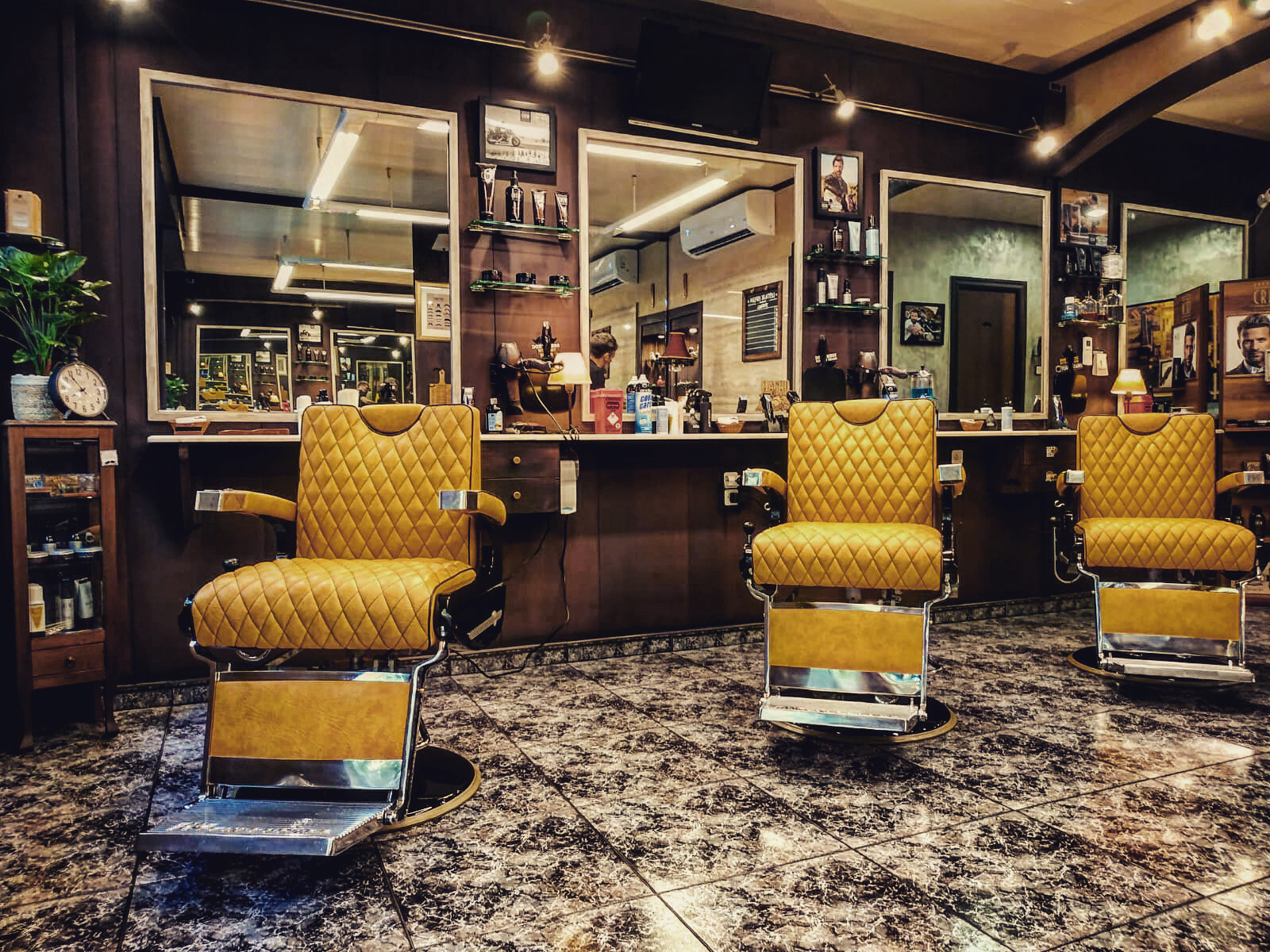 Sala de barbería con tres sillones de piel acolchada color mostaza y reposapiés cromados, paneles de madera oscura, espejos grandes enmarcados y suelo de mármol.