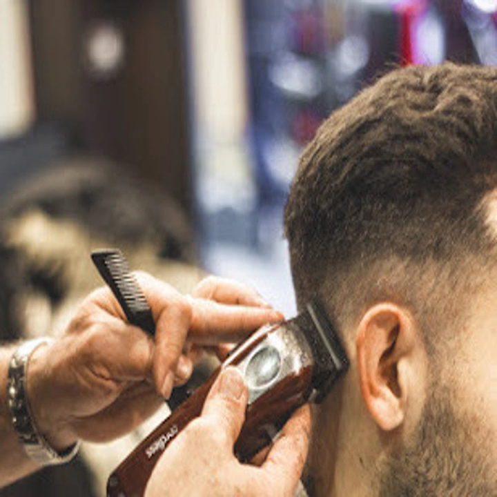Primer plano de las manos de un barbero trabajando un degradado con máquina y peine en la nuca de un cliente.