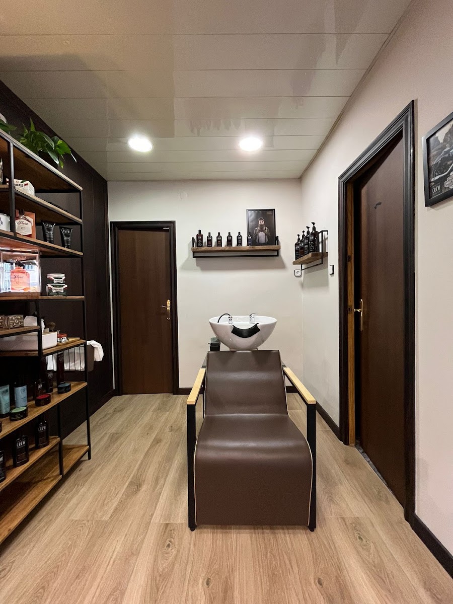 Zona de lavado con un sillón reclinable de piel marrón, lavacabezas blanco, suelo de madera y estanterías de producto.