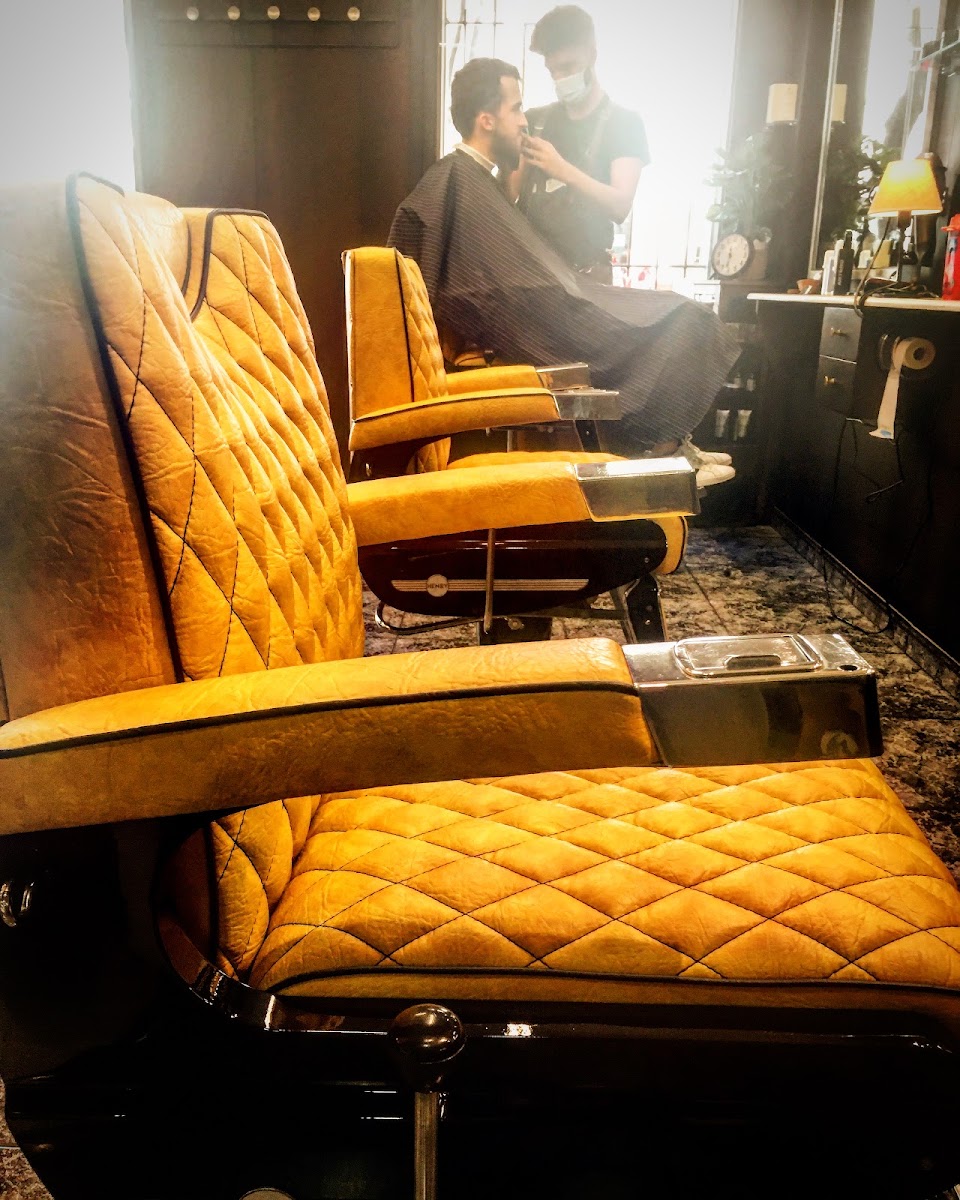 Sillón de barbero de piel mostaza en primer plano; detrás, un barbero recorta la barba a un cliente sentado.