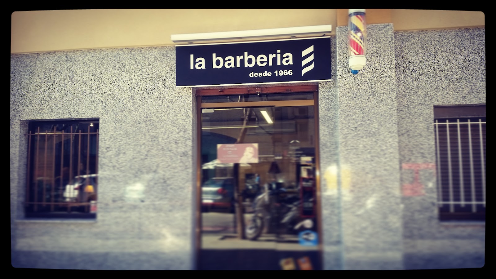 Fachada de La Barbería desde 1966: rótulo azul oscuro con el nombre de la barbería y un logotipo de rayas diagonales, junto a un poste de barbero rojo, blanco y azul.